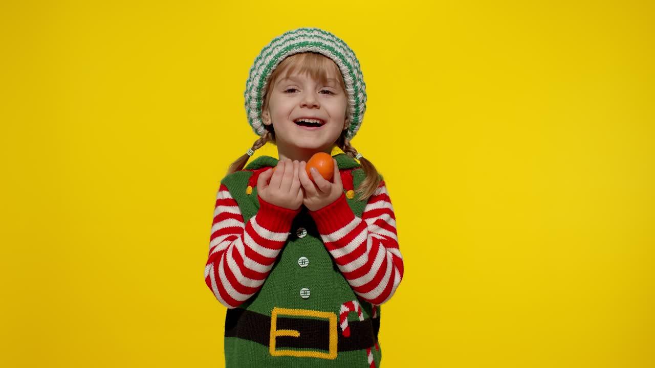 Kid girl in Christmas elf Santa helper costume holding mandarin fruits. New Year holiday celebration