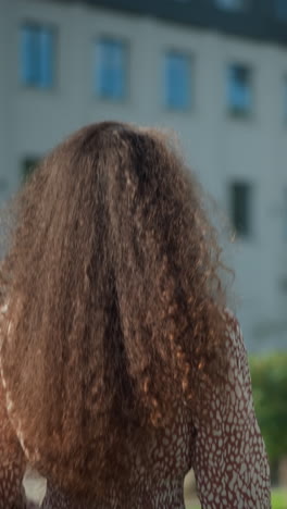 vista trasera de una mujer joven con ropa de época caminando al aire libre, ajustando su cabello rizado, la escena presenta un fondo borroso con edificios modernos, vegetación exuberante y un poste de luz