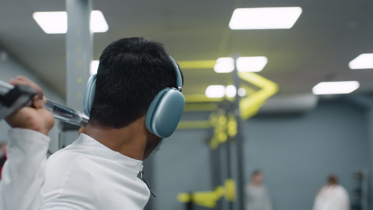 close up of man wearing headphones returning barbell to rack after squat workout in spacious gym with bright ceiling lights and exercise equipment in background