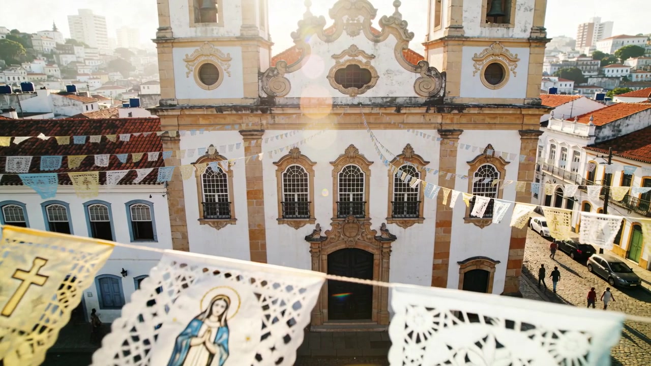 Drone che si allontana rivelando la facciata di una chiesa barocca in una piazza collinare, con papel picado svolazzante.