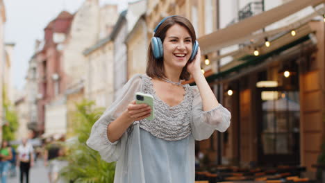 happy young woman in wireless headphones choosing, listening music dancing outdoors city street