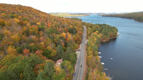 aerial view of dark vehicle on road in colorful autumn landscape, vivid forest by lake sunapee, new hampshire usa, tilt up tracking drone shot