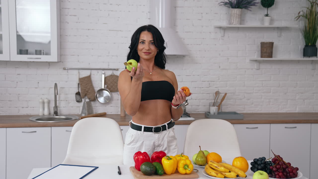 Beautiful woman in her kitchen holding fruit in both hands. Lady wearing black top and white jeans looks at fruit in her hands and then offers a peach and an apple.