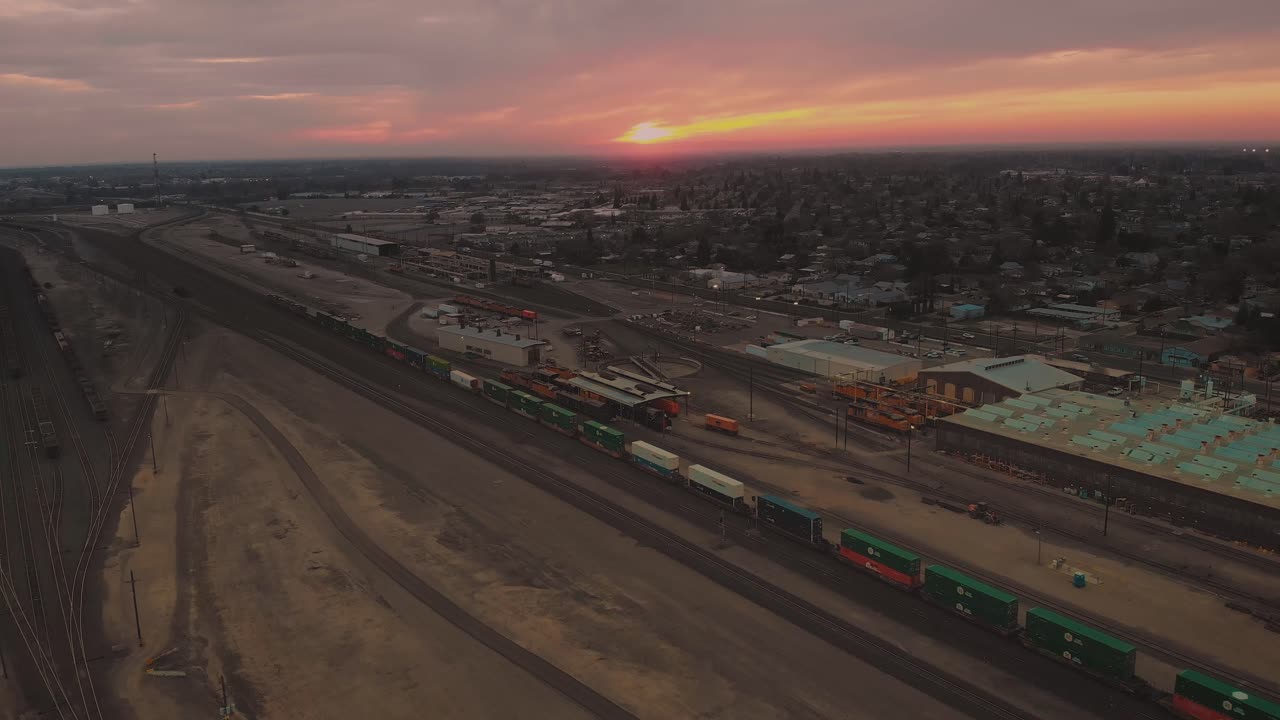 alejando la vista aérea cinematográfica de la parte superior de la ciudad del centro de roseville, california, con edificios industriales, tiendas y trenes de carga que pasan por el ferrocarril con una puesta de sol rosa anaranjada vívida en el fondo