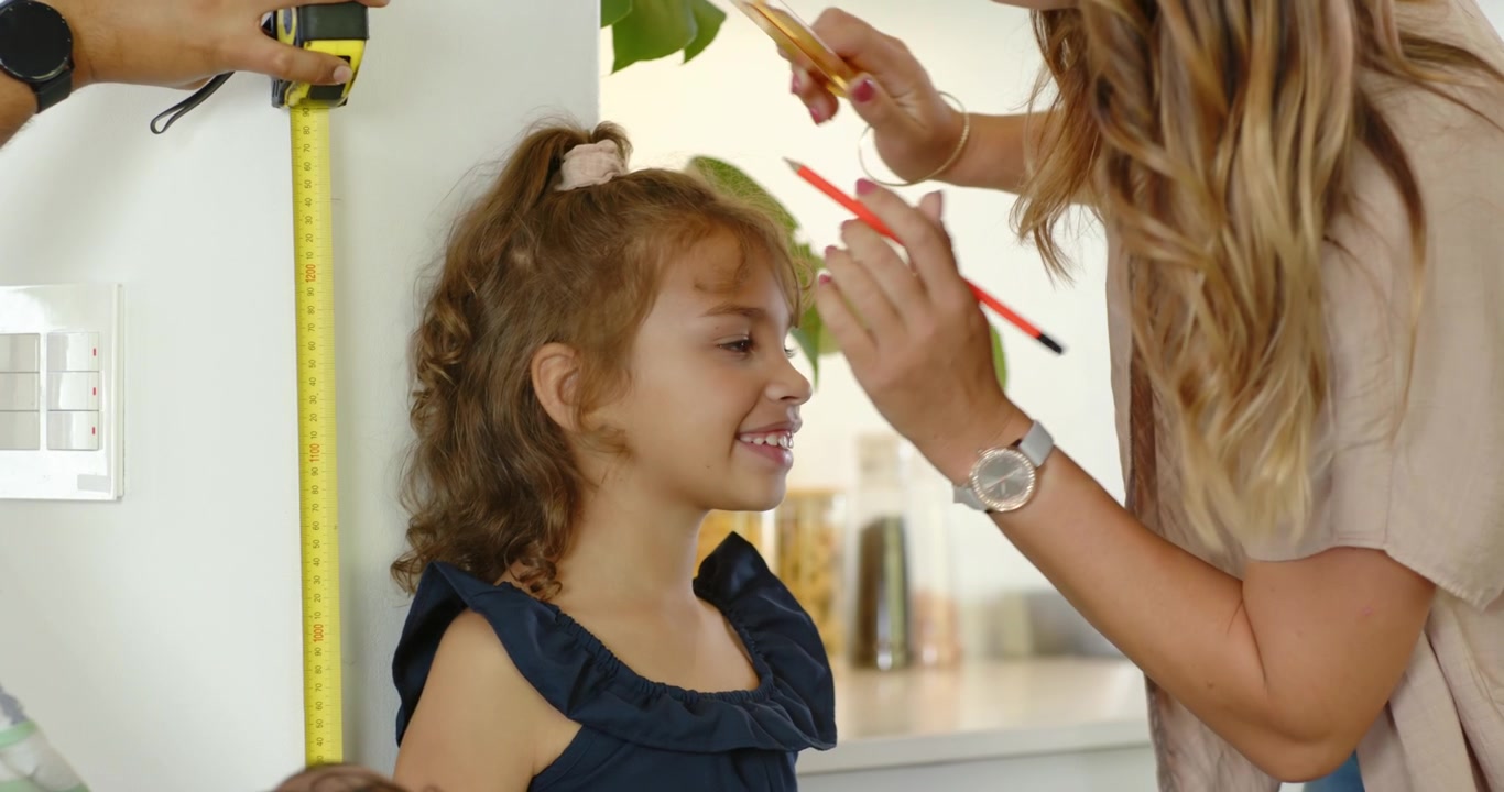 Family measuring daughter in kitchen with yellow tape measure, pencil marking head, tracking growth