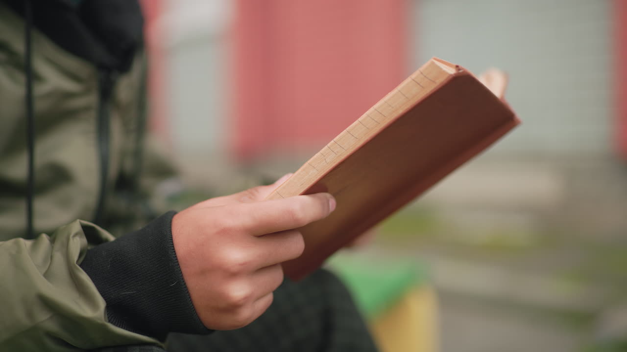 Close up side view of student in green jacket holding book while studying outdoors, flipping to new page with concentration and focus, showing pursuit of knowledge in blurred background
