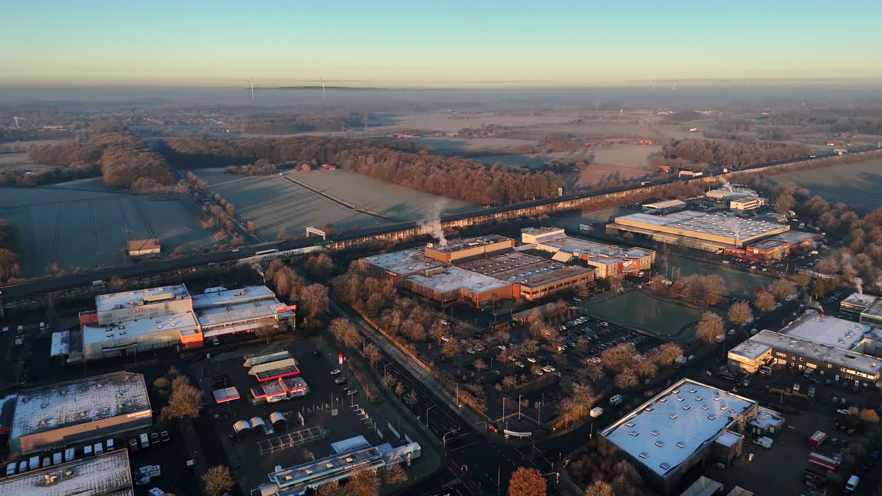 Frozen rooftops of company and factory during golden sunrise in industrial area. Traffic on highway and snowy farm fields in distance. Aerial wide shot.