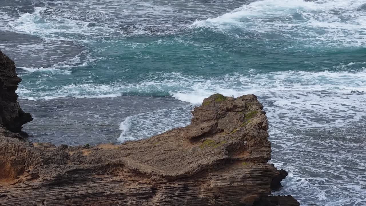 Potenti onde oceaniche si infrangono contro una frastagliata formazione rocciosa marrone sotto condizioni di luce diurna nuvolose