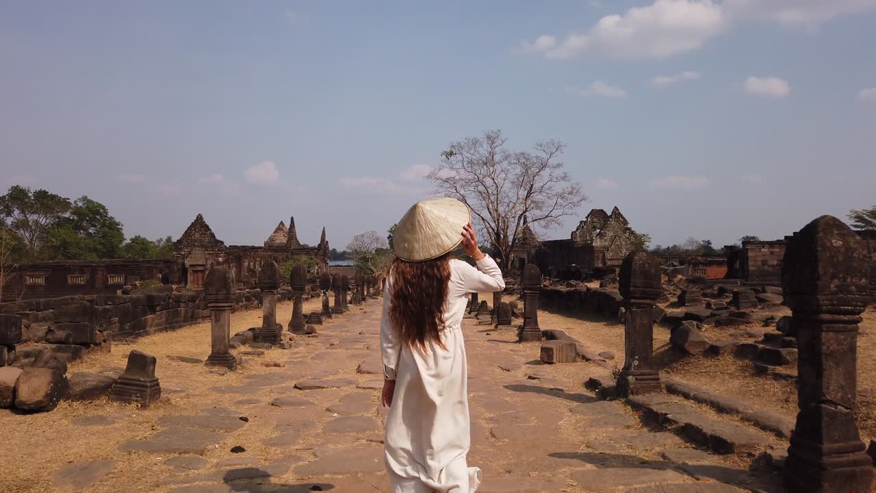Young white woman traveler in long white dress and vietnamese hat walking in Vat Phou – ruined Khmer Hindu Temple. Champassak, Laos, Asia. Sunny day. Ancient culture, religion, ruins. Slow motion.
