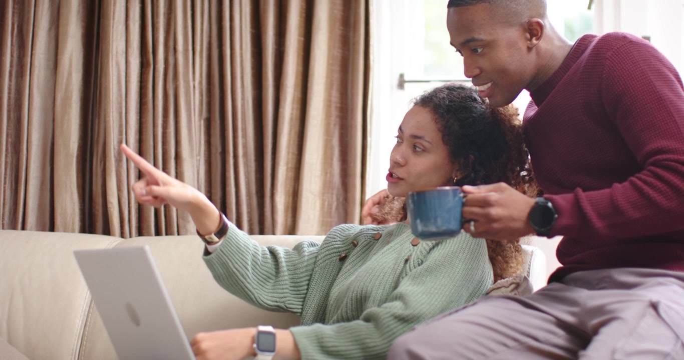 Coppia afroamericana sul divano, la donna digita sul laptop coinvolgendo l'uomo con una tazza blu.