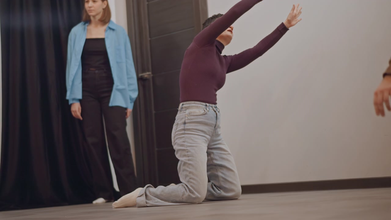 Taneční soubor nacvičující emocionální práci na zemi ve studiu, tanečníci se plazí a natahují, pozorovatel stojí, choreograf vede skupinu, syrová zranitelnost, intenzivní dýchání, výrazná gesta.