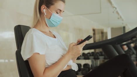 Young-sporty-woman-with-face-mask-uses-a-smartphone-in-the-gym.-Close-up.