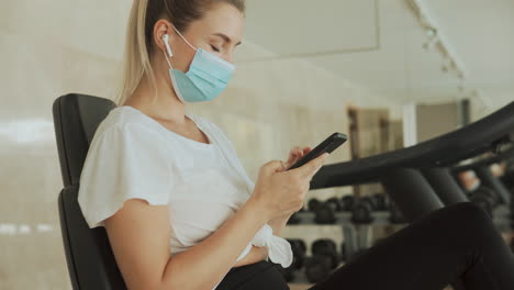 Una-Joven-Deportista-Con-Mascarilla-Utiliza-Un-Teléfono-Inteligente-En-El-Gimnasio.-De-Cerca.