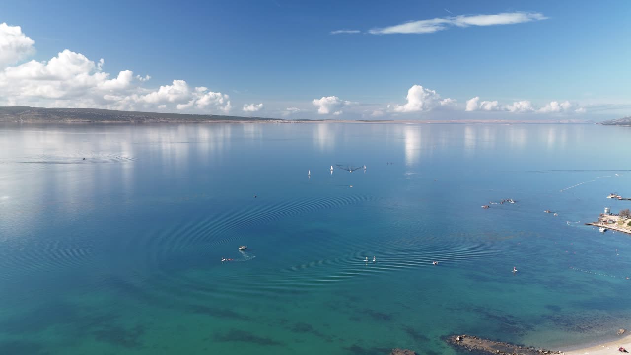 Aerial drone footage over the blue Adriatic Sea in Starigrad-Paklenica, Croatia. Dinghy boats and SUPs glide across calm water with a stunning coastal panorama and white clouds in the distance