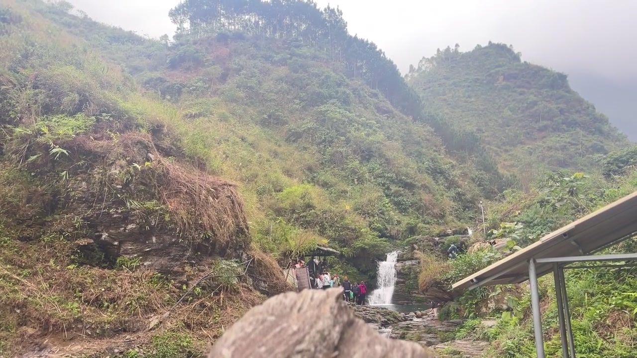 Et jevnt stigende avsløringsbilde går fra bak en stor stein for å vise frem en frodig grønn fjellside, fjerne tåkete topper og en liten rennende foss i Đồng Văn Karstplatået