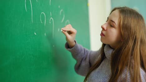 student girl at school. side view of a schoolgirl standing near the blackboard and writing with a white chalk. teenager writes question marks on a board.