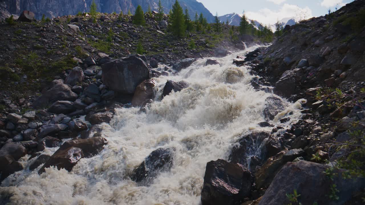 Un fiume impetuoso si infrange su rocce frastagliate, circondato da un aspro terreno montuoso. L'acqua schiuma bianca mentre precipita attraverso gole rocciose. Pini verdi punteggiano i pendii.