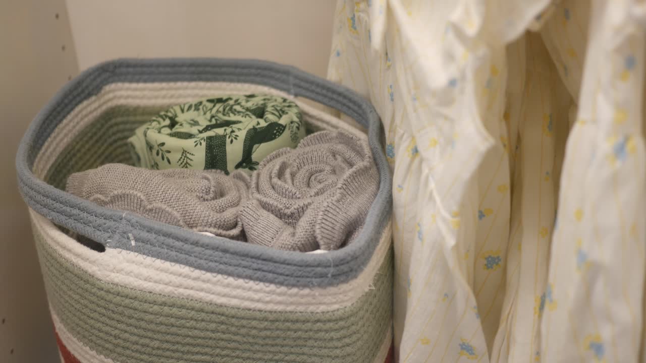A close-up of a woven storage basket with folded blankets and a dress hanging in a closet