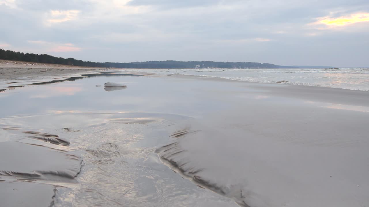 Static side view of waves rolling towards empty pristine beach with white illuminated sand. Forest line in the horizon with foaming waves reaching the shore. Water flowing on wet sand surface, Estonia