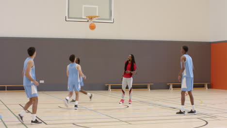 multiracial male basketball players throwing ball into basket with coach in gym
