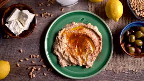 Amplio-Deslizamiento-Hacia-La-Derecha-Que-Muestra-Un-Plato-Plano-De-Hummus-Con-Queso-Feta,-Aceitunas,-Vaso-De-Ouzo,-Limones,-Piñones-Sobre-Una-Mesa-De-Madera-Rústica