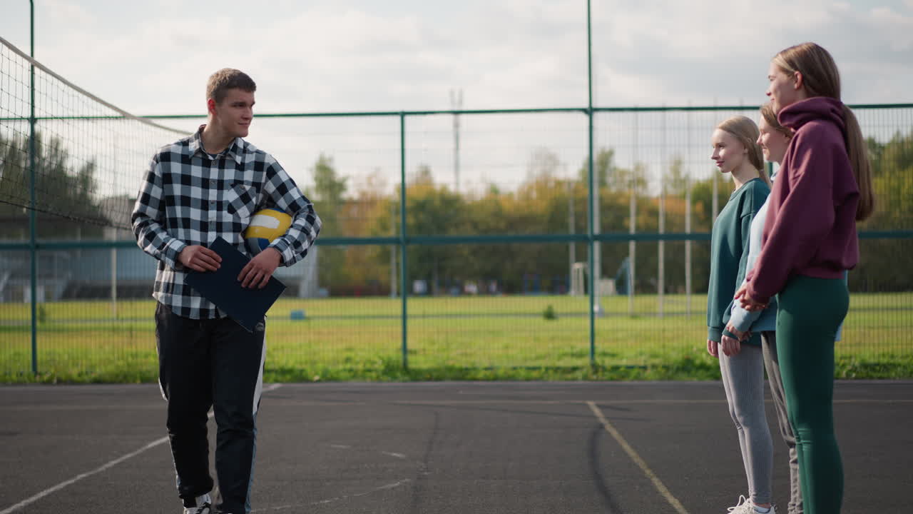 тренер по волейболу идет к студентам с мячом под рукой и записью в руке, студенты тепло улыбаются, когда наблюдают инструктора на открытой площадке