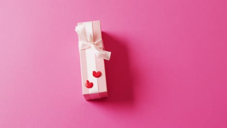 Video-of-pale-pink-gift-box-with-white-bow-and-red-hearts,-on-pink-background-with-copy-space