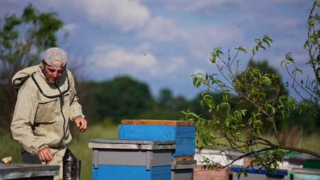 caucasian adult man working in the rural apiary locating in the nature backdrop. beekeeper checking up the frames using tools.
