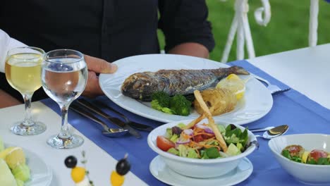 waiter serving a plate of fish to a man guest in a outdoor restaurant, cropped close up view of the food and his hand