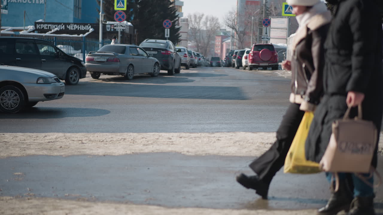winter city street with parked cars and moving traffic, pedestrians in coats and boots passing signs and posts, snow along curb glistening on wet asphalt, urban commute scene at intersection