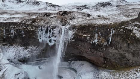 aerial drone shot of seljalandsfoss waterfall in iceland during winter surrounded by snow-covered cliffs, frozen streams, rugged terrain, icy slopes, scenic views, people walking near base.