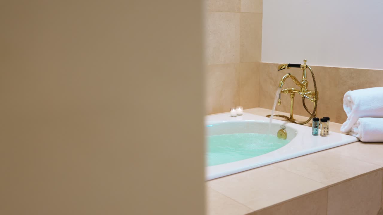 Luxurious and romantic bubble bath is filled by a golden tap while a bottle of champagne is waiting in a wine cooler and the towels are neatly rolled up. Close up panning shot