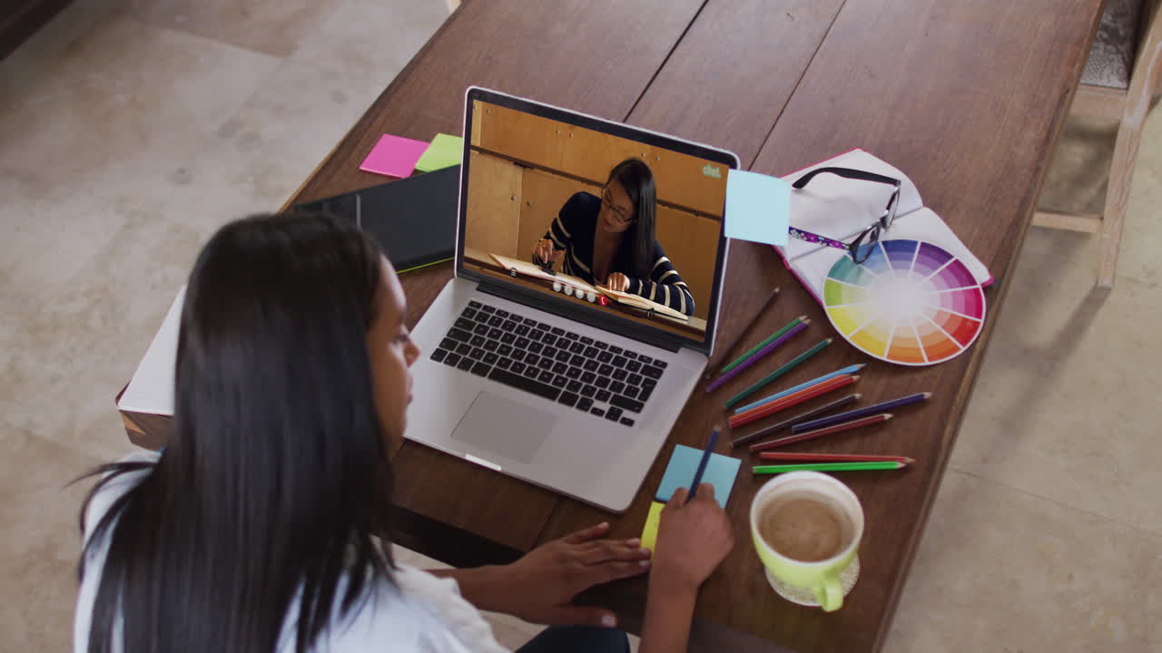 Profesora caucásica usando una laptop en videollamada con una estudiante, tomando apuntes.