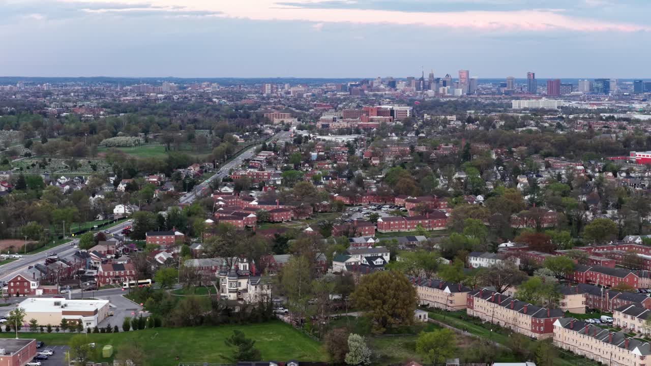 Panorama aéreo de un barrio suburbano de Baltimore con casas adosadas de ladrillo y edificios de apartamentos, que se extiende hacia el horizonte del centro bajo la suave luz nublada de la mañana y un exuberante entorno verde. Toma amplia