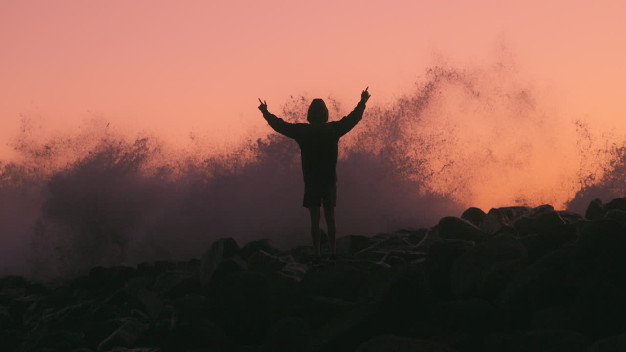 el adolescente levanta las manos cuando la ola choca contra el muelle causando un gran chorro de agua en el fondo al atardecer