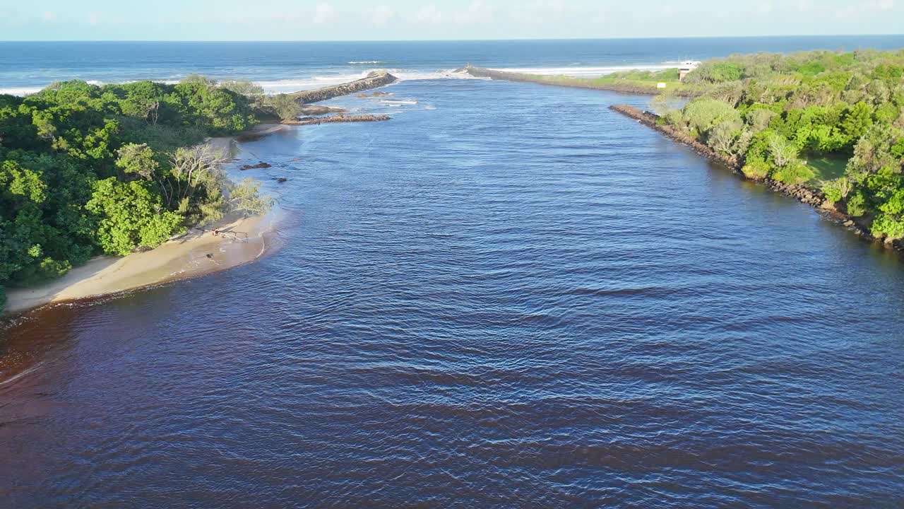 Drone captures serene river flowing into the ocean, surrounded by lush greenery and rocky banks, under soft sunset lighting