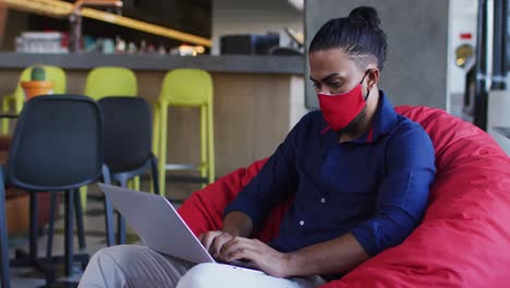 African-american-man-wearing-face-mask-sitting-in-cafe-using-laptop