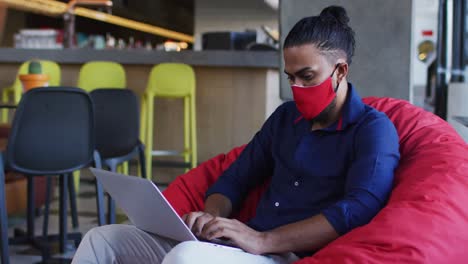hombre afroamericano con máscara facial sentado en una cafetería usando una computadora portátil