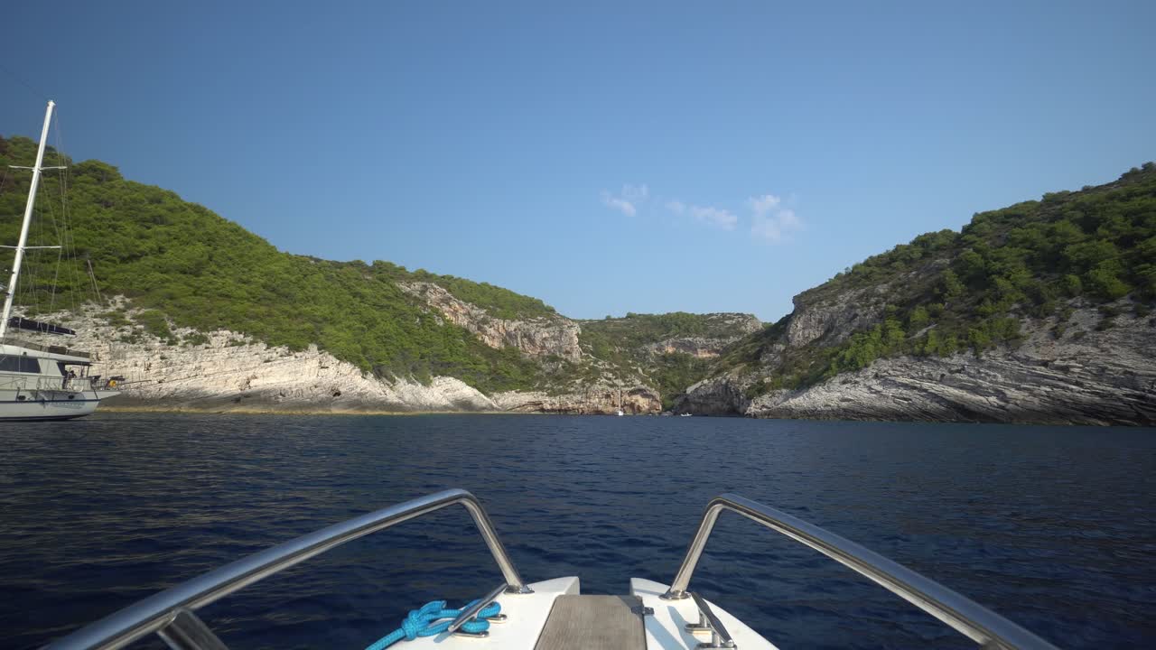 proa de un barco de recreo a toda velocidad a través del agua azul hacia una costa rocosa más allá de un yate de vela