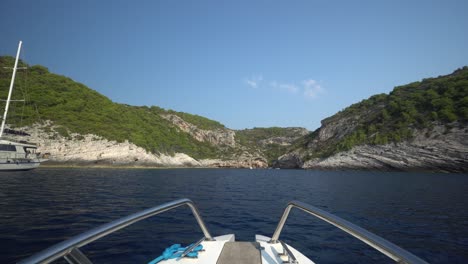 Bow-of-recreational-boat-speeding-across-blue-water-toward-a-rocky-shoreline-past-a-sailing-yacht