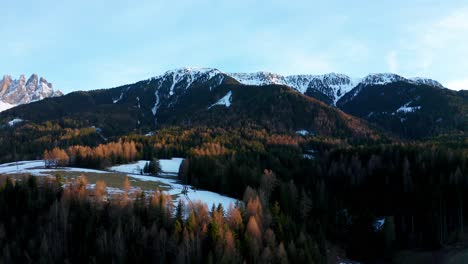 Drone-view-of-a-forest-down-the-Dolomites-mountains-in-Italy-at-sunset-with-sun,-snow,-peaks,-landscape,-light-reflections-and-blue-sky