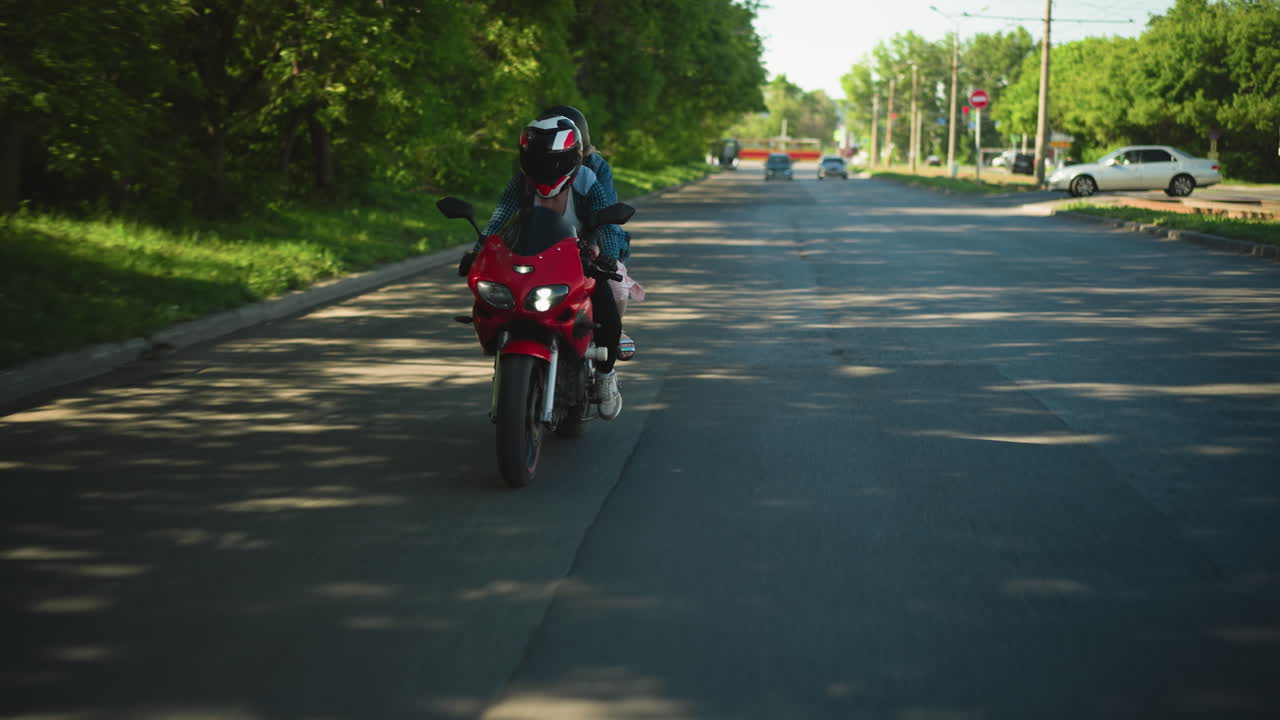 dos mujeres montan una bicicleta eléctrica roja a lo largo de una carretera asfaltada, con una vista borrosa de dos coches que se acercan desde la distancia y un coche estacionado cerca de una curva en u, la carretera está sombreada por árboles