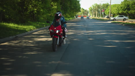 two women ride a red power bike along a tarred road, with a blurred view of two cars approaching from the distance and a parked car near a u-turn, the road is shaded by trees