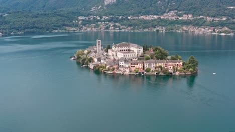 Isola-San-Giulio-En-El-Tranquilo-Lago-Orta,-Italia,-Con-Un-Barco-A-Motor-Pasando-Y-Exuberantes-Colinas-Al-Fondo,-Vista-Aérea