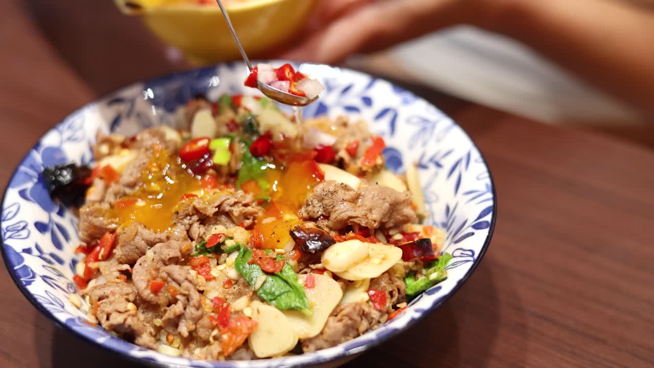 Hand pours fish sauce onto Thai beef, garlic, and chili rice bowl under warm lighting