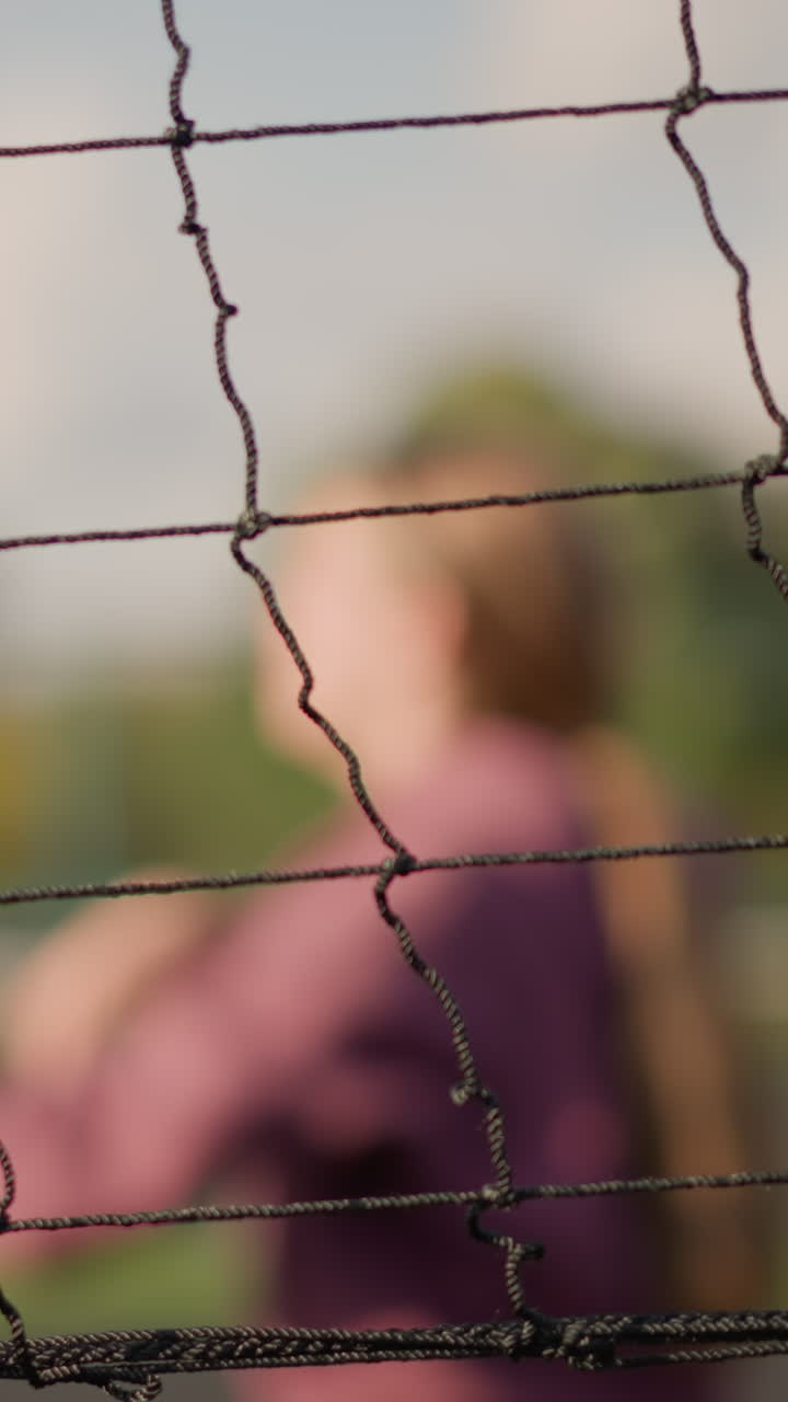 primer plano de una red de voleibol con fondo borroso que muestra a una mujer de color marrón jugando al voleibol con otra persona, la luz del sol y la vegetación crean un resplandor dorado