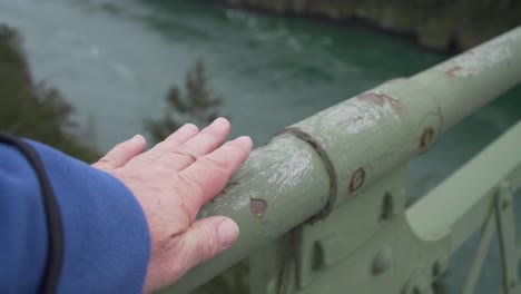 person's hand running down railing of bridge with green fast flowing river below