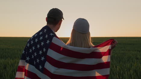 young couple with american flag on their shoulders looking forward in wheat field at sunset