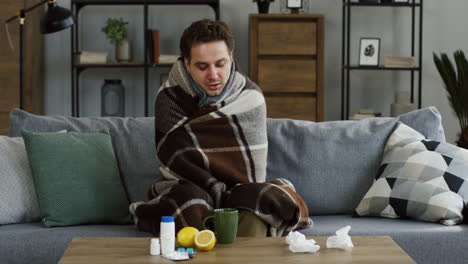 Young-Unwell-Man-In-The-Warm-Scarf-Shivering-As-He-Having-A-High-Temperature-And-A-Cold,-Sitting-On-The-Couch-In-The-Living-Room