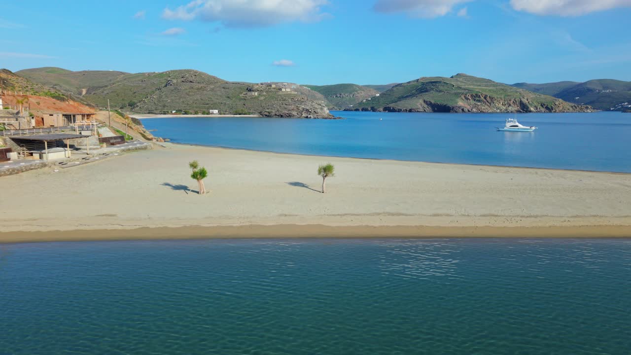 Aerial establishing overview of Kolona Beach with anchored boats, sandbar, and hilly terrain in background, telephoto tracking right